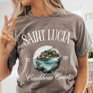 May include: A grey t-shirt with a white graphic design that says "Saint Lucia Est. 1979 Caribbean Coast". The graphic features a watercolor illustration of a tropical island with palm trees and a beach.