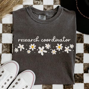 May include: A charcoal gray t-shirt with the words "research coordinator" in white script, adorned with a floral design of white daisies with yellow centers. White canvas sneakers and a black hat are also visible.