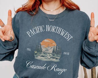 May include: A blue-grey sweatshirt with the text "Pacific Northwest Cascade Range" in white script. The graphic features a mountain scene with a sunset. The person in the image is wearing blue jeans and making a peace sign.