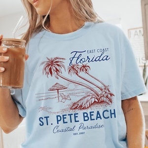 May include: Light blue t-shirt with a graphic of palm trees and a beach scene. The text "East Coast Florida" and "St. Pete Beach Coastal Paradise Est. 1957" is printed on the shirt.