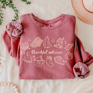 May include: A dusty rose crewneck sweatshirt featuring a white line-art graphic of autumn-themed items including a blanket, pumpkin, drink, turkey, mug, sock, and pie, with the text "thankful optician" centred in the middle of the design.