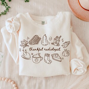 Puede incluir: Una sudadera de manga larga color crema con un gráfico de líneas marrones de elementos otoñales que incluyen un pavo, una calabaza, un pastel y una taza de café, con el texto "thankful radiologist" impreso en el centro del diseño.