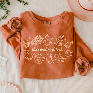 May include: A burnt orange long-sleeve crewneck sweatshirt featuring a white line-art graphic of autumn-themed items including a pumpkin, a turkey, a pie slice, a mug, and a blanket, with the text "thankful rad tech" printed in the center.