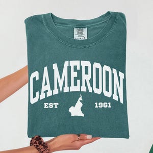 May include: A teal-colored t-shirt with the word "CAMEROON" in large white block letters. Below the word are the words "EST" and "1961", along with a white silhouette of the country. The shirt is held up by a person.
