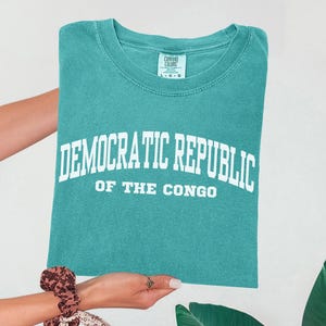 May include: Teal-colored t-shirt with the words "DEMOCRATIC REPUBLIC OF THE CONGO" in white block letters. The shirt is held up by a person. The shirt has a crew neck and a tag that says "COMFORT COLORS".