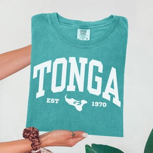May include: A teal short-sleeved t-shirt featuring the white text TONGA in a collegiate font, with the text EST 1970 and a white silhouette map of the island nation positioned between the years. The shirt has a Comfort Colors brand neck label.