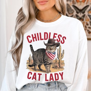 May include: White long-sleeve sweatshirt with the text "Childless Cat Lady" in red. A cartoon cat wearing a cowboy hat and bandana is standing in front of cacti.