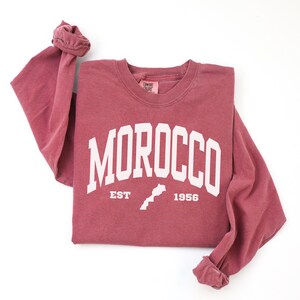 May include: A long-sleeve crewneck sweatshirt in a muted berry red color. The front features white screen-printed text that reads MOROCCO in an arched font, with EST 1956 and a small map outline of the country positioned below the text.