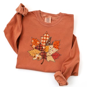 May include: A long-sleeve crewneck sweatshirt in a muted terracotta orange color. The center features a large maple leaf graphic composed of various patchwork patterns including plaid, floral, and polka dot prints in shades of orange, red, and cream.
