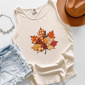 Puede incluir: Una camiseta sin mangas de color crema con un gráfico central de una hoja de arce compuesta por varios patrones de patchwork en tonos naranja, rojo, marrón y amarillo. La prenda incluye una etiqueta de la marca Comfort Colors en el cuello.