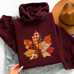 May include: A deep maroon hooded sweatshirt featuring a large, central graphic of a maple leaf composed of various patchwork-style fabric patterns in shades of orange, red, yellow, and brown, with visible stitching details throughout the leaf design.