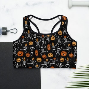 May include: A black racerback sports bra featuring an all-over print of white skeletons and orange jack-o'-lantern pumpkins. The skeletons are depicted in various dancing poses, while the pumpkins display different carved facial expressions.