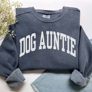 May include: A dark gray crewneck sweatshirt featuring the white text DOG AUNTIE in a bold, arched collegiate font across the chest. The garment has long sleeves with ribbed cuffs and a visible interior fleece lining, with a Comfort Colors brand label.