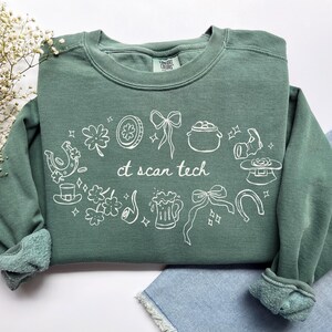 May include: A sage green crewneck sweatshirt featuring white line art graphics of St. Patrick's Day symbols including shamrocks, horseshoes, a pot of gold, a beer mug, and bows. The text "ct scan tech" is printed in the center in white script.
