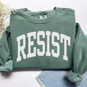 May include: A sage green sweatshirt with the word "RESIST" printed in bold, white, collegiate-style letters across the front. The sweatshirt has a crew neck and ribbed cuffs. The garment is folded on a white surface with a sprig of white flowers.