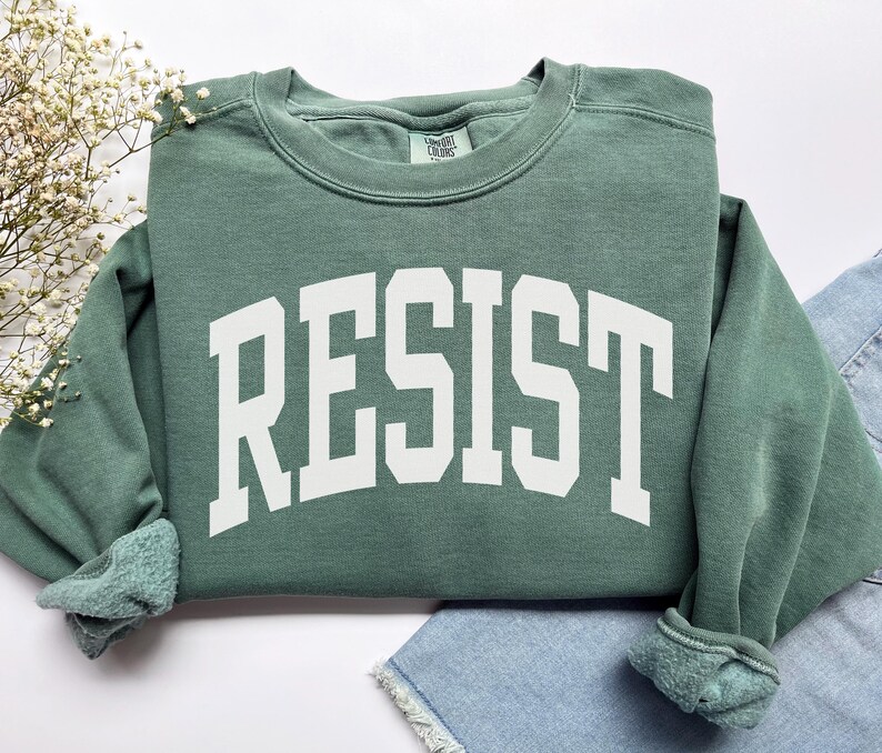 May include: A sage green sweatshirt with the word "RESIST" printed in bold, white, collegiate-style letters across the front. The sweatshirt has a crew neck and ribbed cuffs. The garment is folded on a white surface with a sprig of white flowers.