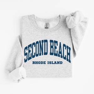 May include: A light gray sweatshirt with the words "SECOND BEACH" in navy blue arched lettering. Below the text is "RHODE ISLAND" in a smaller navy blue font. The sleeves are rolled up.