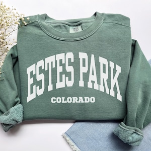 May include: A sage green sweatshirt with the text "ESTES PARK COLORADO" in white block letters. The sweatshirt has a crew neck and ribbed cuffs and hem. The garment is made of a soft, comfortable material.