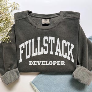 Op de afbeelding: Een donkergrijze sweatshirt met de tekst "FULLSTACK DEVELOPER" in grote witte letters. De crew neck sweatshirt heeft manchetten en is opgevouwen op een spijkerbroek. Het kledingstuk is een casual stijl.