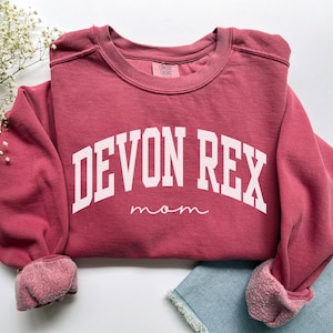 May include: A maroon sweatshirt with white block lettering that reads "DEVON REX" above the word "mom" in a cursive font. The sweatshirt has a crew neck and ribbed cuffs. The sleeves are rolled up.