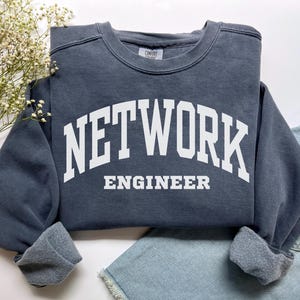 Puede incluir: Una sudadera gris oscuro con las palabras "NETWORK ENGINEER" impresas en blanco, arqueadas sobre el pecho. La sudadera tiene cuello redondo y puños. La prenda está doblada sobre una superficie blanca con una ramita de flores blancas y unos shorts vaqueros azules.