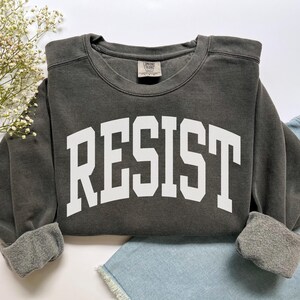 May include: A dark gray sweatshirt with the word "RESIST" printed in large, white, block letters across the front. The sweatshirt has a crew neck and cuffed sleeves. The garment is folded on top of a pair of blue denim shorts.