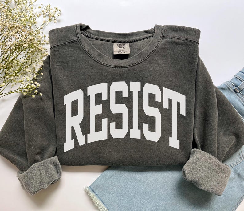 May include: A dark gray sweatshirt with the word "RESIST" printed in large, white, block letters across the front. The sweatshirt has a crew neck and cuffed sleeves. The garment is folded on top of a pair of blue denim shorts.
