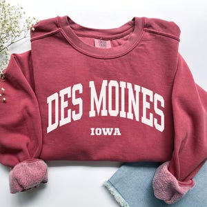 May include: A maroon sweatshirt with the text "DES MOINES IOWA" in white block letters. The sweatshirt has a crew neck and cuffed sleeves. The fabric appears soft and comfortable.