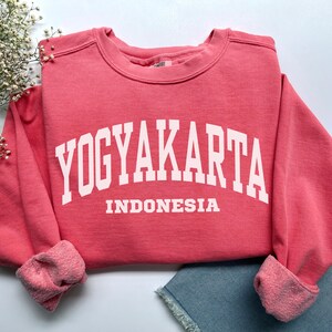 Puede incluir: Una sudadera de color coral con la palabra "YOGYAKARTA" en letras blancas grandes, con "INDONESIA" debajo. La sudadera tiene cuello redondo y puños acanalados. La prenda está doblada, con una parte de unos pantalones cortos vaqueros azules visibles.