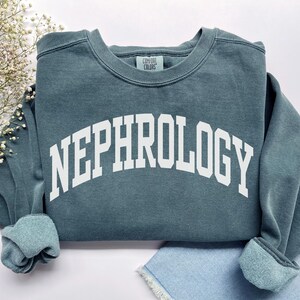 Puede incluir: Una sudadera de color verde azulado con la palabra "NEPHROLOGY" impresa en letras blancas en el pecho. La sudadera tiene cuello redondo y puños acanalados. La etiqueta de la marca Comfort Colors es visible.