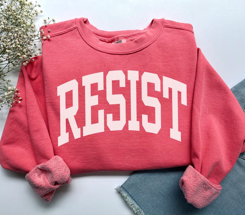 May include: A coral-colored sweatshirt with the word "RESIST" printed in large, white, block letters across the front. The sweatshirt has a crew neck and long sleeves with ribbed cuffs. The garment is folded, with a portion of denim shorts visible.