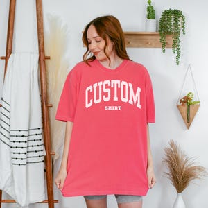 以下が含まれることがあります： 胸に白いブロック体で「CUSTOM」の文字がアーチ状に、その下に「SHIRT」とプリントされた、コーラル色のTシャツ。 ゆったりとしたフィット感で、半袖です。