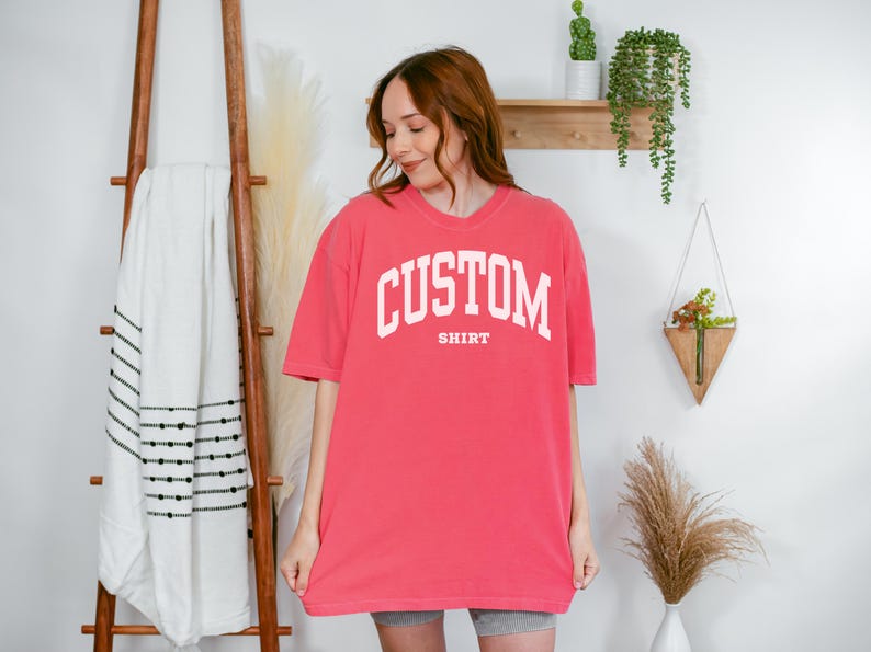 以下が含まれることがあります： 胸に白いブロック体で「CUSTOM」の文字がアーチ状に、その下に「SHIRT」とプリントされた、コーラル色のTシャツ。 ゆったりとしたフィット感で、半袖です。