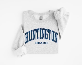 Könnte beinhalten: Ein hellgraues Sweatshirt mit dem Text "HUNTINGTON BEACH" in marineblauer, bogenförmiger Schrift. Das Sweatshirt hat einen Rundhalsausschnitt und lange Ärmel, wobei die Ärmel hochgekrempelt sind. Das Kleidungsstück liegt flach auf einer weißen Oberfläche.