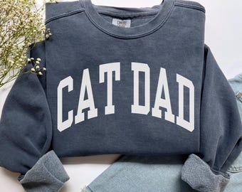 Może przedstawiać: Ciemnoniebieska bluza z białym napisem "CAT DAD" pogrubioną, łukowatą czcionką. Bluza ma okrągły dekolt i mankiety. Ubranie jest prezentowane na białej powierzchni z gałązką białych kwiatów i parą niebieskich szortów dżinsowych.