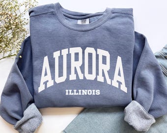 May include: A slate blue crewneck sweatshirt featuring the word AURORA in large white arched block letters with the word ILLINOIS printed in smaller white block letters underneath. The garment has a ribbed collar and long sleeves with ribbed cuffs.