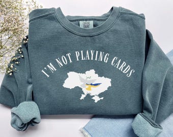 May include: A teal sweatshirt with the text "I'M NOT PLAYING CARDS" in white. The design includes a white outline of Ukraine with a dove and the Ukrainian flag colors. The sweatshirt has a crew neck and ribbed cuffs.