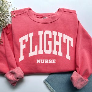 May include: A coral-colored sweatshirt with the words "FLIGHT NURSE" in large, white, block letters across the front. The sweatshirt has a crew neck and ribbed cuffs. The sleeves are cuffed, and the garment is folded.