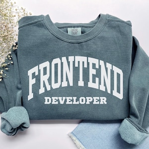 Könnte beinhalten: Ein teal-farbenes Sweatshirt mit dem weißen Aufdruck "FRONTEND DEVELOPER" in Bogenform auf der Vorderseite. Das Sweatshirt hat einen Rundhalsausschnitt und gerippte Bündchen. Das Comfort Colors-Etikett ist sichtbar.