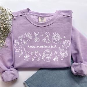 May include: A lilac-colored sweatshirt with white graphic illustrations of Easter-themed elements, including bunnies, ducks, and cupcakes. The text "hoppy anesthesia tech" is printed in the center of the design. The sweatshirt is folded on top of blue denim.