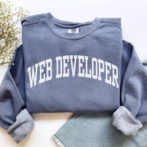 Könnte beinhalten: Ein staubblaues Sweatshirt mit dem Text "WEB DEVELOPER" in fetten, weißen Blockbuchstaben. Das Sweatshirt hat einen Rundhalsausschnitt und gerippte Bündchen. Es ist auf einer hellblauen Jeans gefaltet.