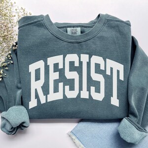 May include: A teal-colored sweatshirt with the word "RESIST" printed in large, white, block letters. The sweatshirt has a crew neck and ribbed cuffs. The Comfort Colors brand tag is visible at the neckline.