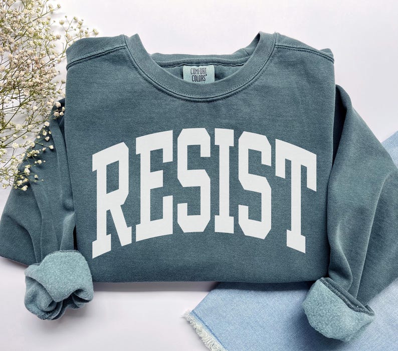 May include: A teal-colored sweatshirt with the word "RESIST" printed in large, white, block letters. The sweatshirt has a crew neck and ribbed cuffs. The Comfort Colors brand tag is visible at the neckline.