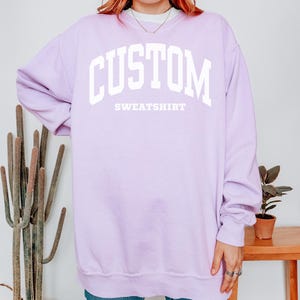 以下が含まれることがあります： 「CUSTOM」の文字が大きく白でプリントされた、ライラック色のスウェットシャツ。「SWEATSHIRT」の文字がその下にあります。クルーネックと長袖のデザインです。モデルは青いジーンズを着用しています。