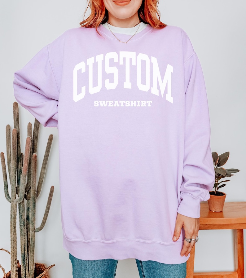 以下が含まれることがあります： 「CUSTOM」の文字が大きく白でプリントされた、ライラック色のスウェットシャツ。「SWEATSHIRT」の文字がその下にあります。クルーネックと長袖のデザインです。モデルは青いジーンズを着用しています。