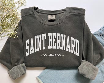 May include: Dark grey sweatshirt featuring the text "SAINT BERNARD" in white block letters and "mom" in script. The crew neck sweatshirt has cuffed sleeves. The garment is made of a soft material.