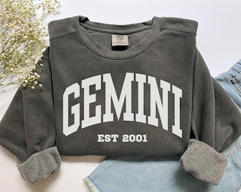 Op de afbeelding: Een antracietgrijze sweater met ronde hals en de witte tekst GEMINI in een groot gebogen lettertype, met de tekst EST 2001 eronder gecentreerd. Het kledingstuk heeft een geribbelde kraag, boorden en zoom, met een Comfort Colors label.