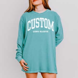 以下が含まれることがあります： ターコイズブルーの長袖シャツ。白い大きなブロック体で「CUSTOM」の文字が「LONG SLEEVE」の上に書かれています。 ゆったりとしたフィット感とクルーネックが特徴です。 テキストはカレッジスタイルのフォントです。