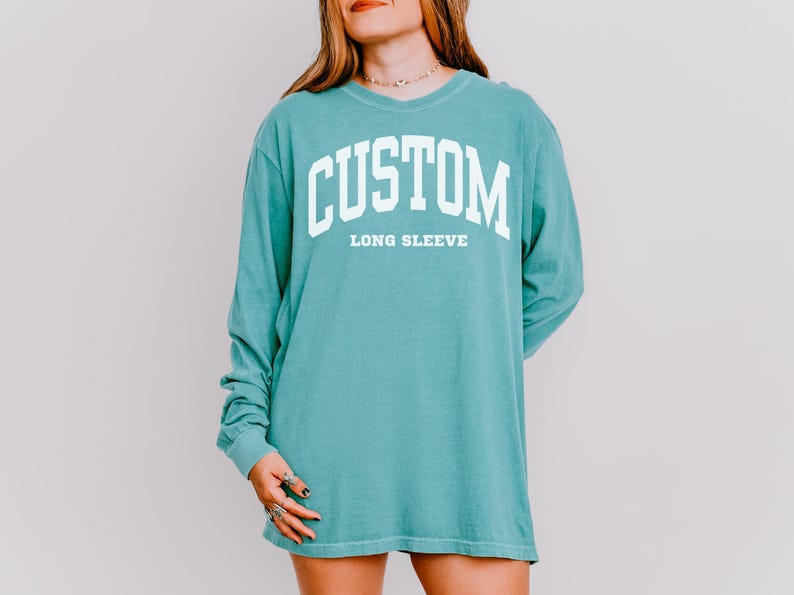 以下が含まれることがあります： ターコイズブルーの長袖シャツ。白い大きなブロック体で「CUSTOM」の文字が「LONG SLEEVE」の上に書かれています。 ゆったりとしたフィット感とクルーネックが特徴です。 テキストはカレッジスタイルのフォントです。