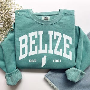 Puede incluir: Sudadera de cuello redondo color turquesa con la palabra "BELIZE" en letras blancas grandes. Debajo, el texto "EST 1981" y un contorno blanco de Belice. La sudadera está doblada sobre unos vaqueros azules.